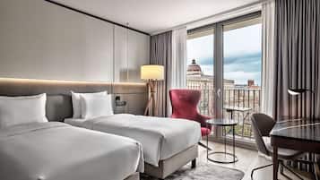 Habitación de hotel con dos camas, un escritorio, una silla, una lámpara y vistas a un paisaje urbano a través de amplios ventanales.