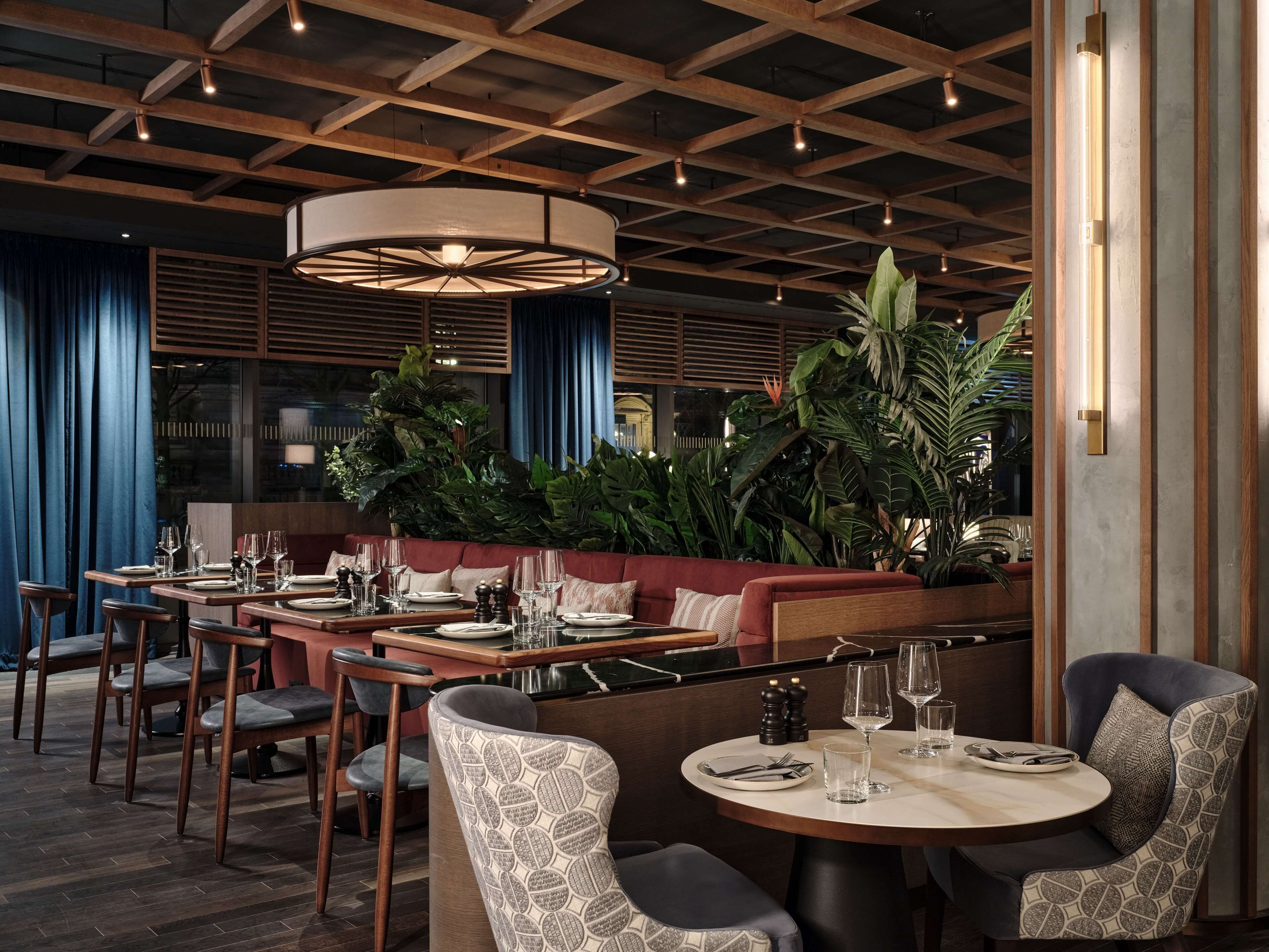 L’intérieur d’un restaurant avec des poutres en bois au plafond, des tables rondes, des sièges rembourrés et de grandes plantes en pot.