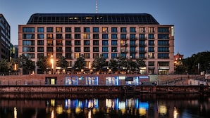 A modern building with a flat roof and multiple windows, reflecting on a calm body of water.
