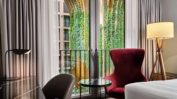 Habitación de hotel con cama, un sillón rojo, una mesita, una lámpara de pie y vistas a zonas verdes a través de una puerta de cristal.
