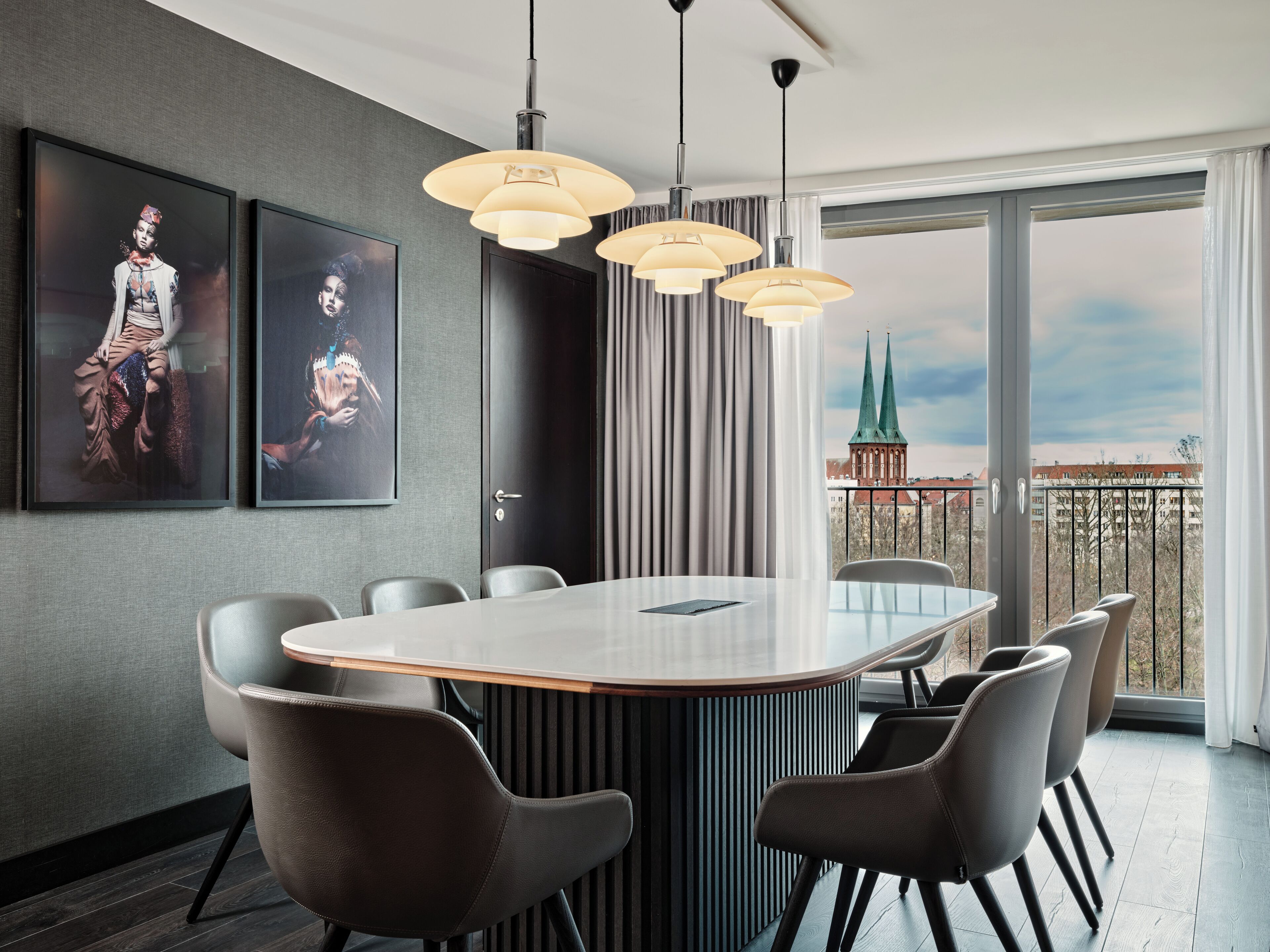 Ein moderner Essbereich mit einem großen Tisch, sechs Stühlen und Pendelleuchten. An der Wand befinden sich zwei gerahmte Kunstwerke und ein Blick auf die Stadt durch ein großes Fenster.