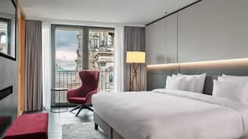 Ett hotellrum med en stor säng, en röd fåtölj, ett litet bord och utsikt över en historisk byggnad.