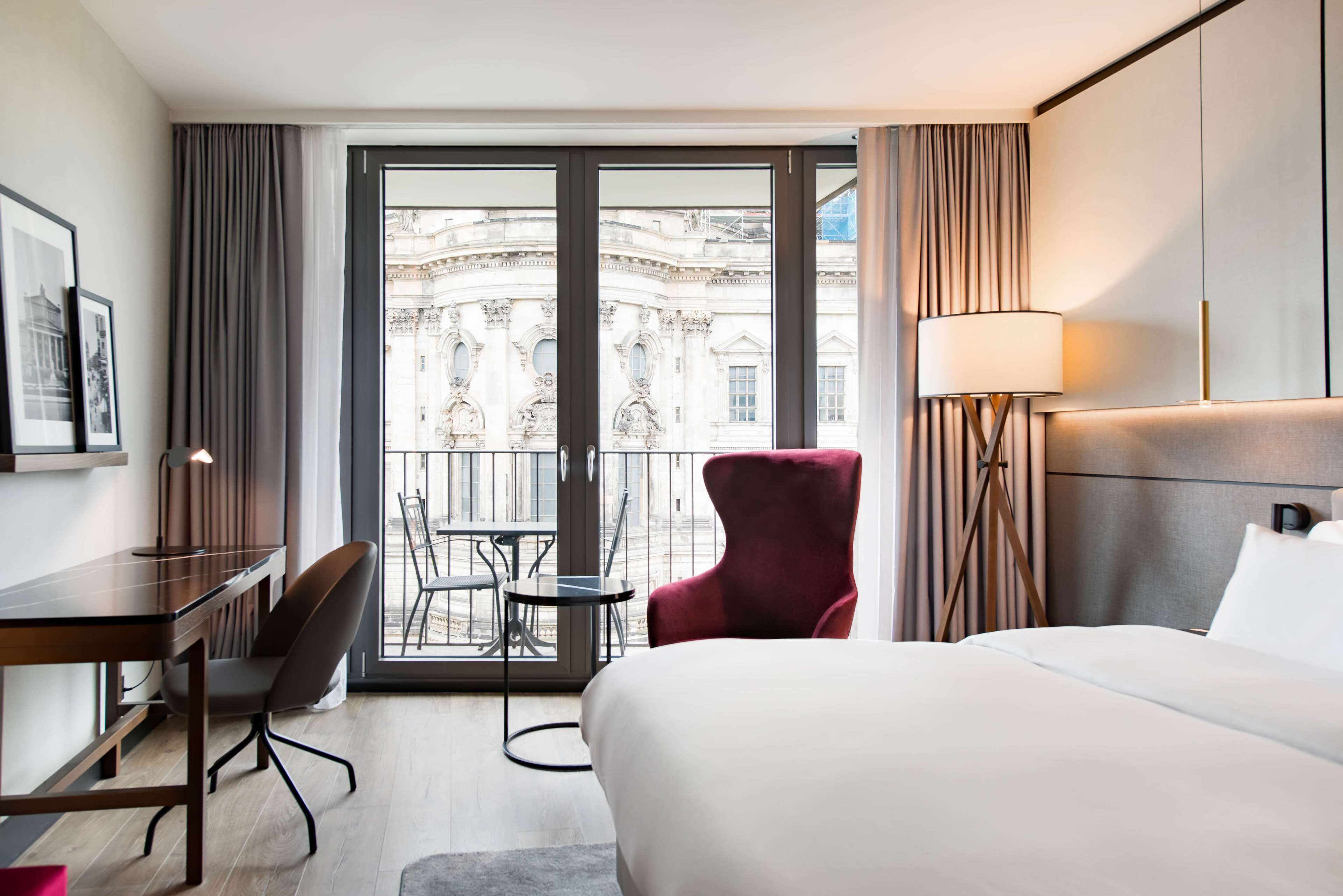 A hotel room with a large bed, a desk, a chair, and a balcony with a view of a historic building.