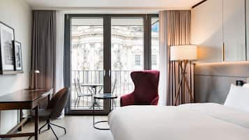 Habitación de hotel con una cama grande, un escritorio, una silla y un balcón con vistas a un edificio histórico.
