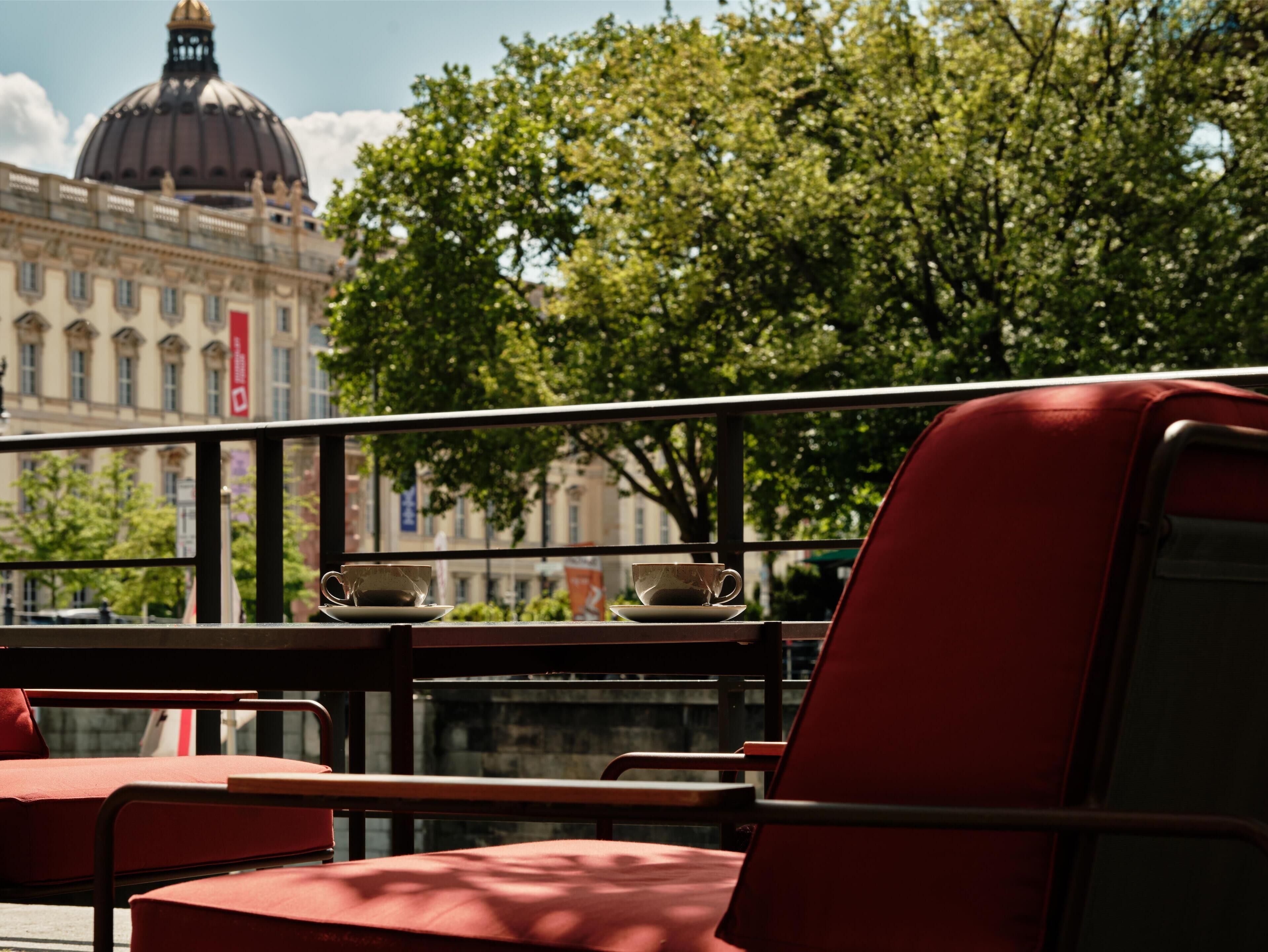 Un café con sillas y mesas rojas, vista de un edificio con cúpula y árboles al fondo.