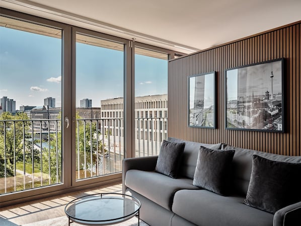 A modern living room with a grey sofa, a glass coffee table, and a city view through large windows.