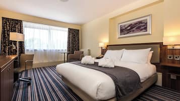 A hotel room with a large bed, a desk, a chair, a window with curtains, a painting on the wall, and a carpeted floor.