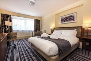 A hotel room with a large bed, a desk, a chair, a window with curtains, a painting on the wall, and a carpeted floor. - Holiday Inn Warrington by IHG (Warrington)