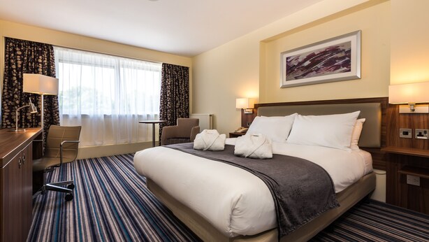 A hotel room with a large bed, a desk, a chair, and a window with curtains. - Holiday Inn Warrington by IHG (Warrington)