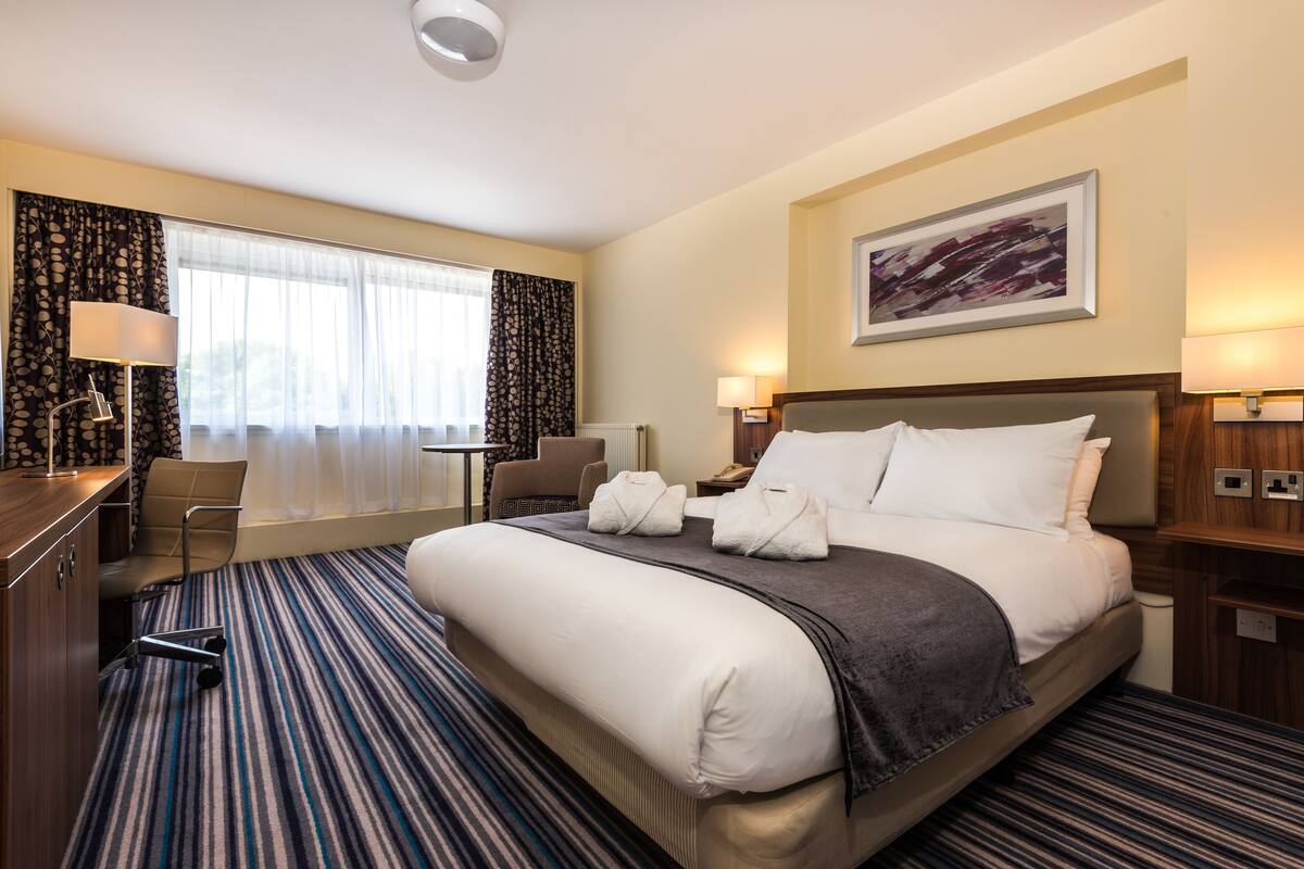 A hotel room with a large bed, a desk, a chair, and a window with curtains.