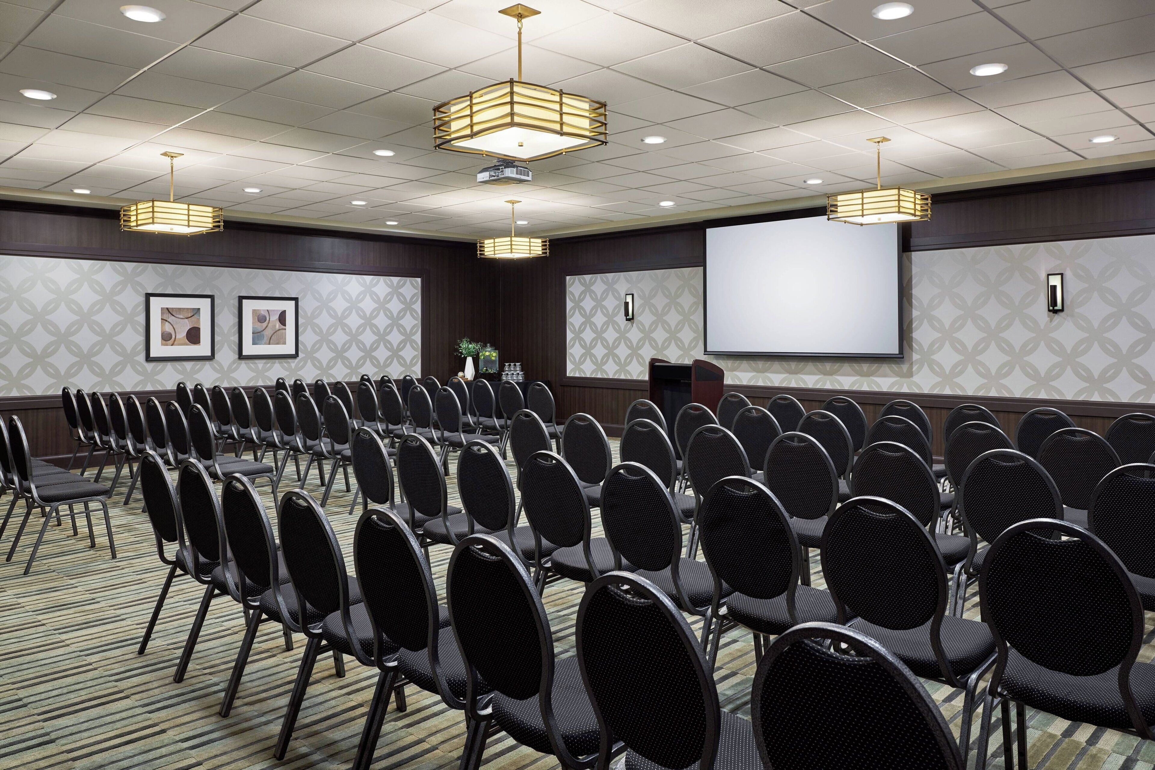 A conference room with rows of chairs, a projector screen, and a large screen on the wall.