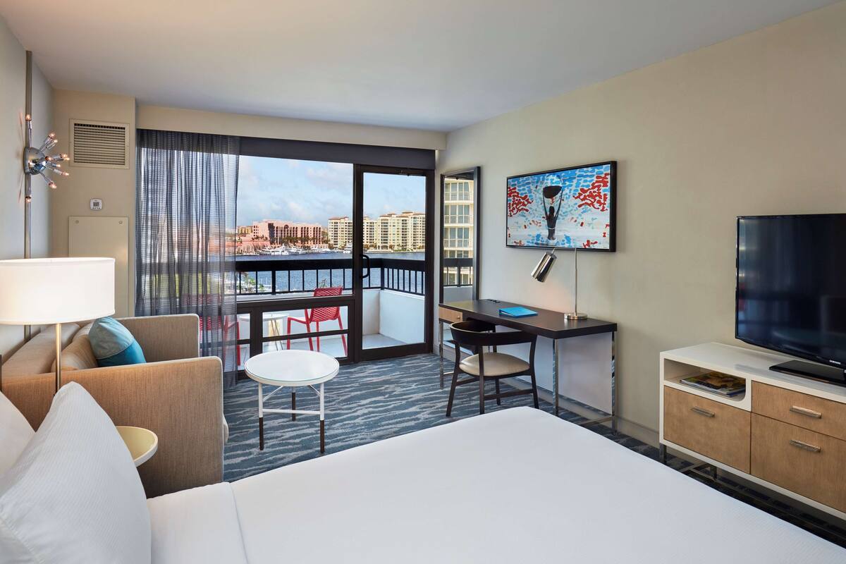 A hotel room with a bed, a desk, a chair, a TV, and a balcony with a view of buildings.