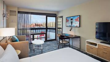 Habitación de hotel con cama, escritorio, silla, televisor y balcón con vistas a edificios.