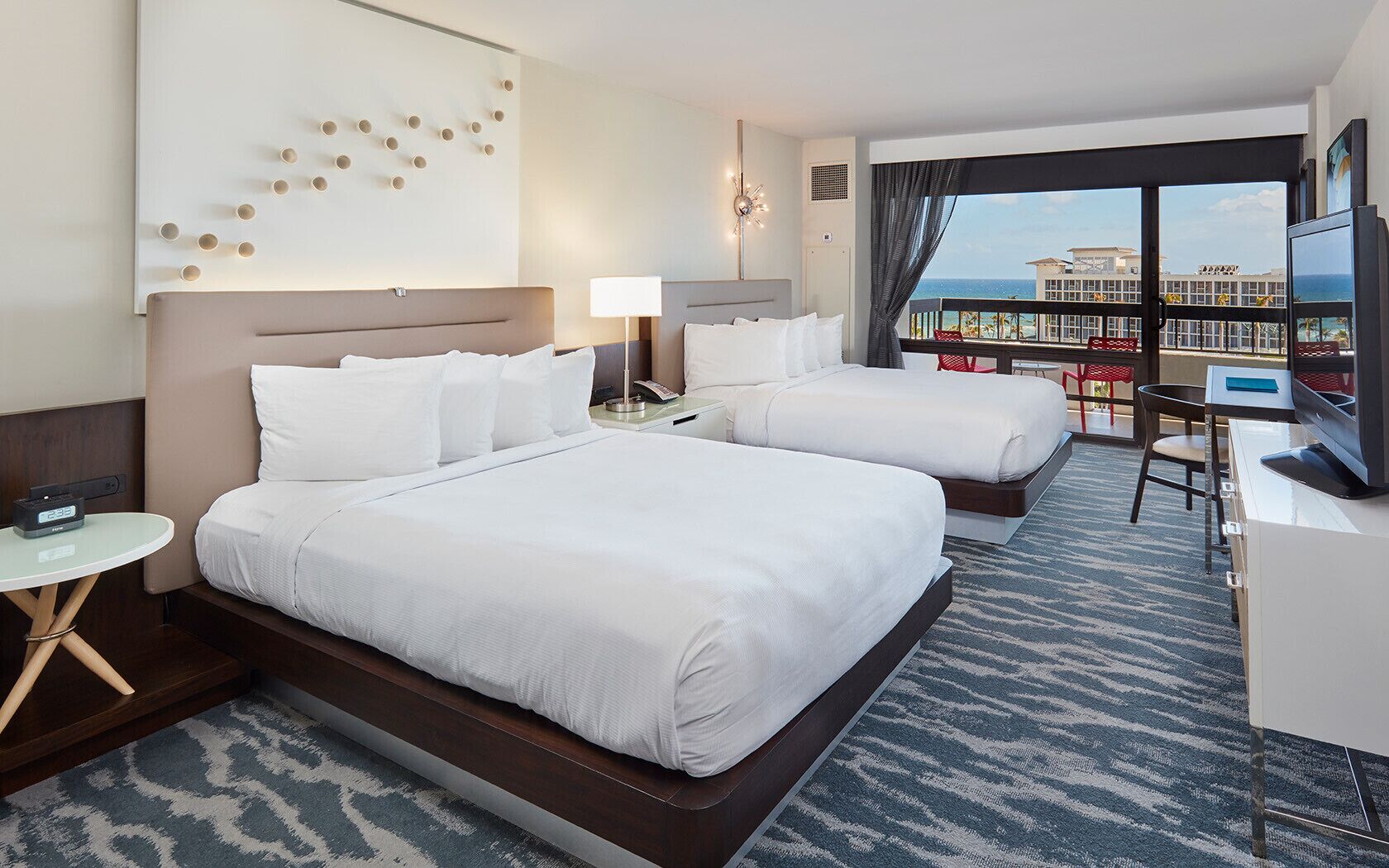 A hotel room with two beds, a TV, a desk, and a view of the ocean.