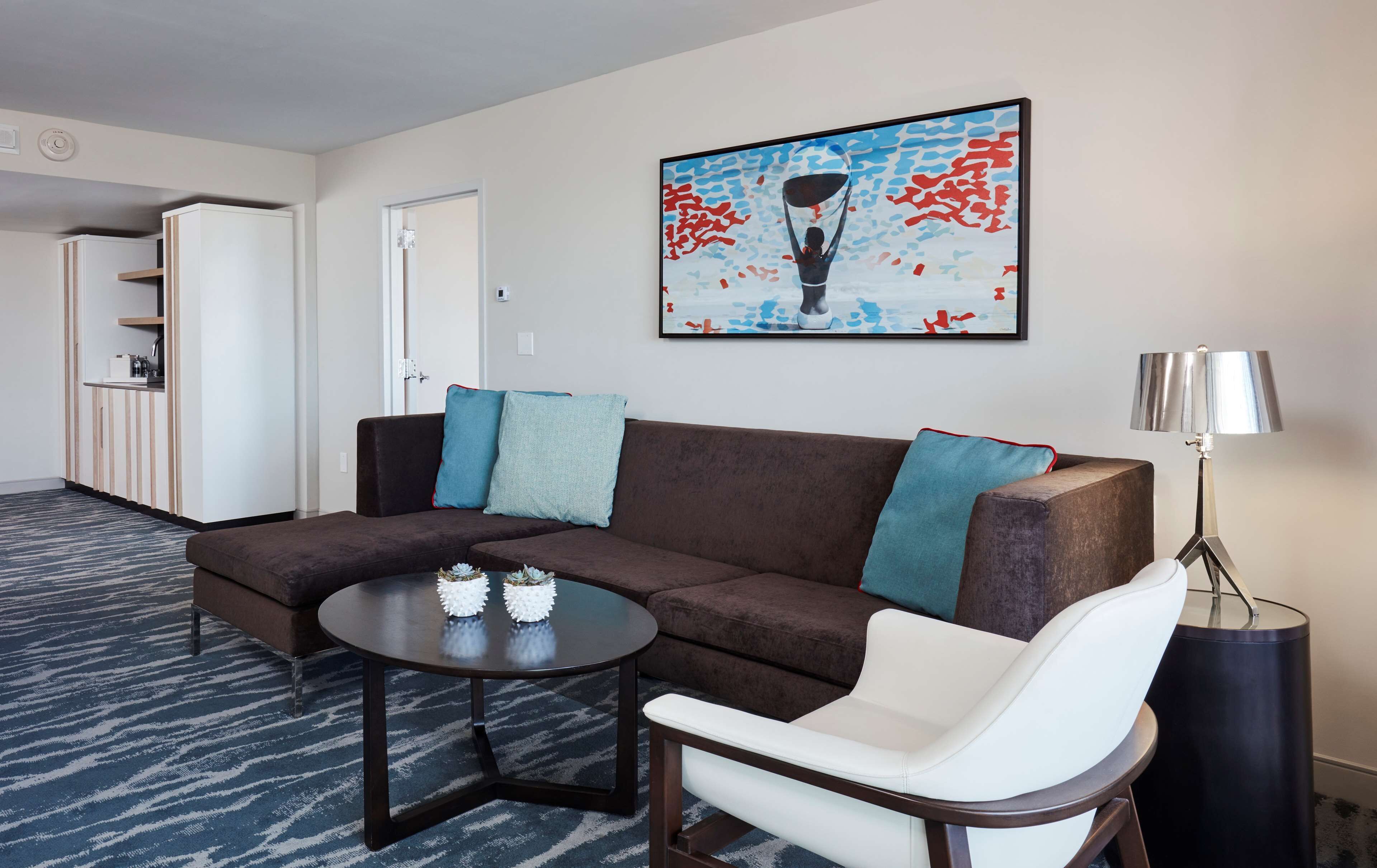 Un salón moderno con un sofá marrón, un sillón blanco y una pequeña mesa de centro. Hay un cuadro en la pared y una lámpara en una mesita auxiliar.