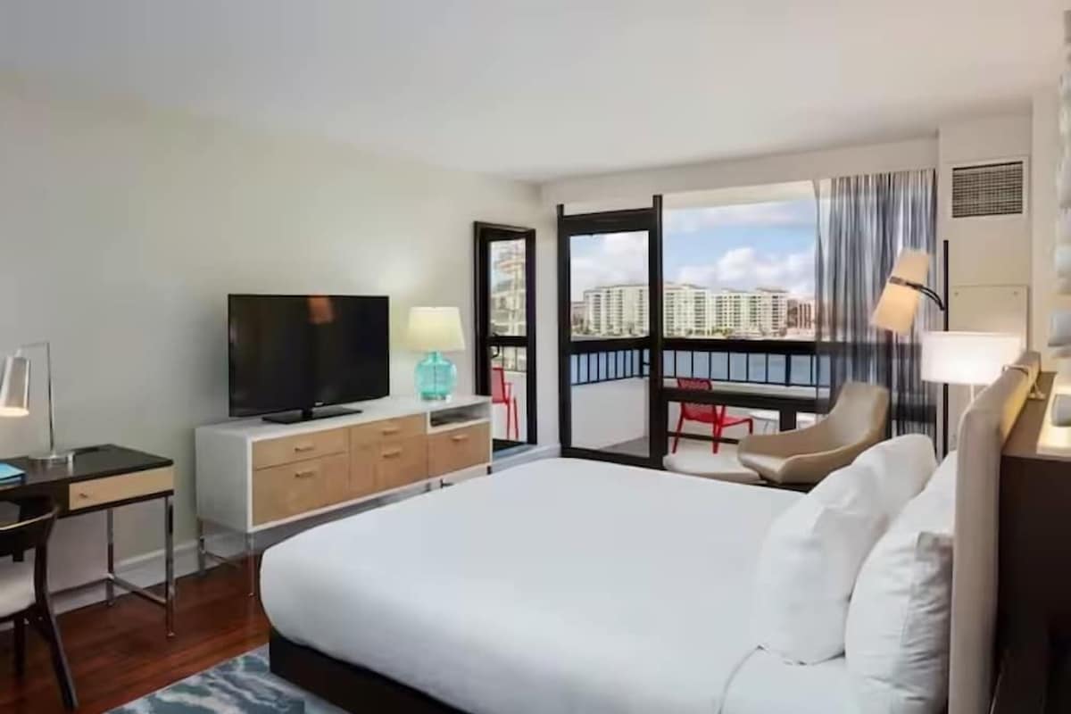 A hotel room with a bed, a desk with a TV, a chair, a balcony with a view of buildings.