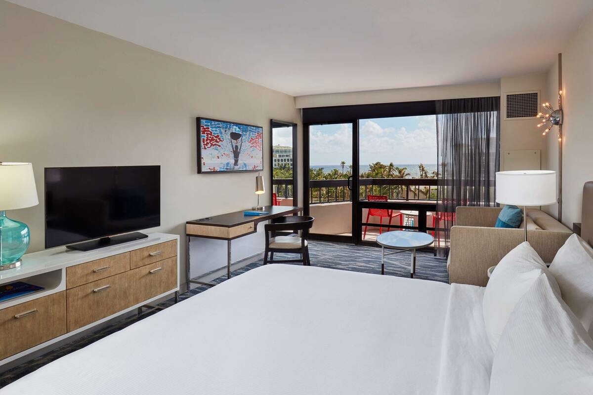 A hotel room with a large bed, a desk with a TV, a balcony with a view of the ocean, and a painting on the wall.