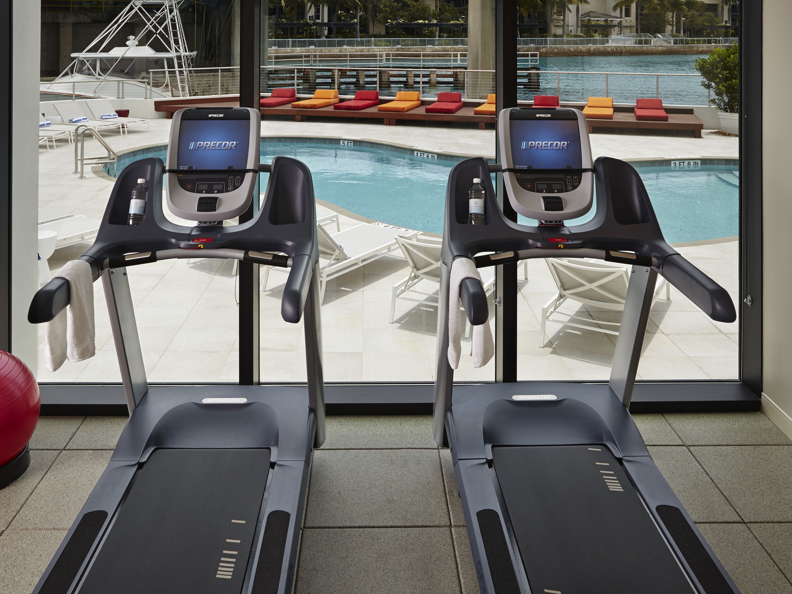Dos cintas de correr con pantallas Precor, una piscina y zona de estar exterior con cojines rojo y naranja.
