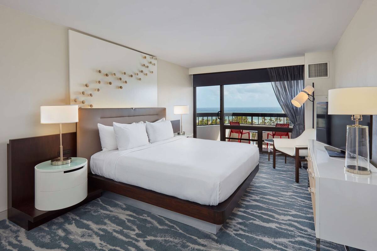 A modern hotel room with a large bed, a desk, and a balcony with a view.