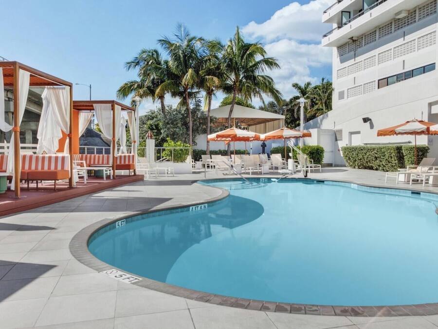 Una piscina con sillas de descanso y sombrillas, rodeada de palmeras y un edificio moderno.