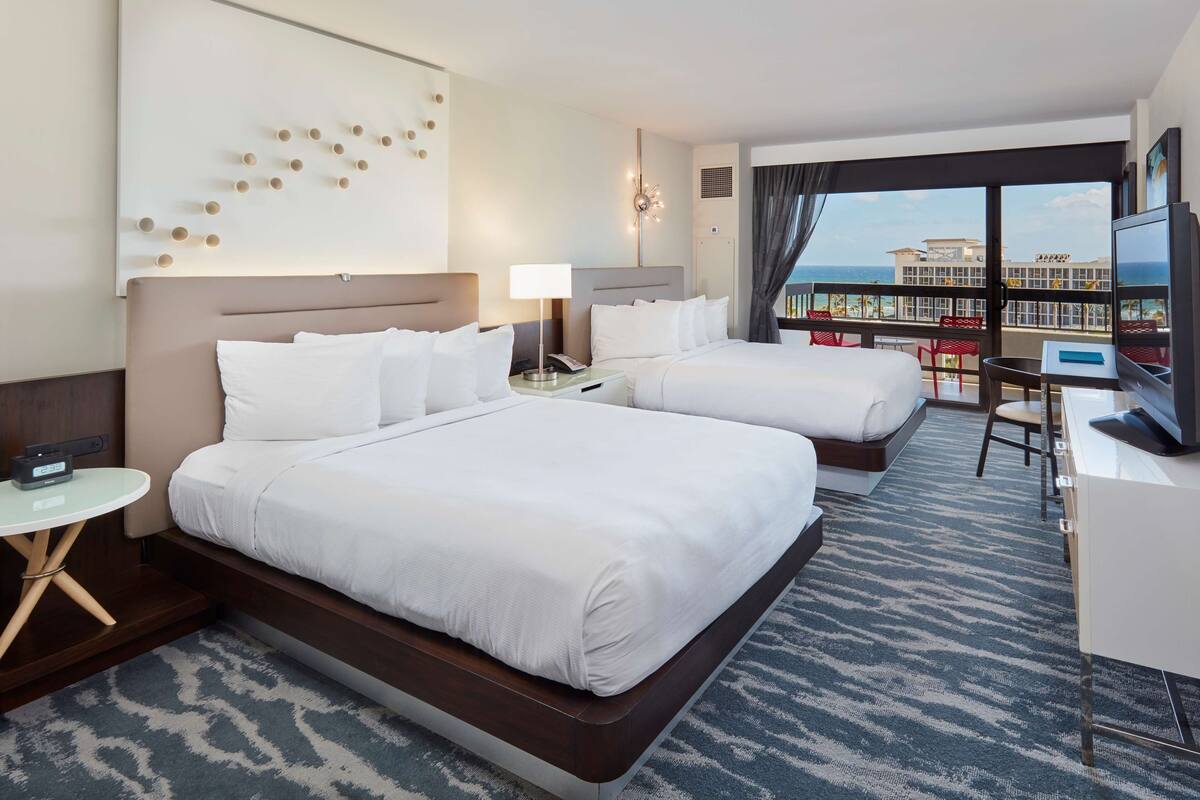 A hotel room with two beds, a desk, a TV, and a view of the city.