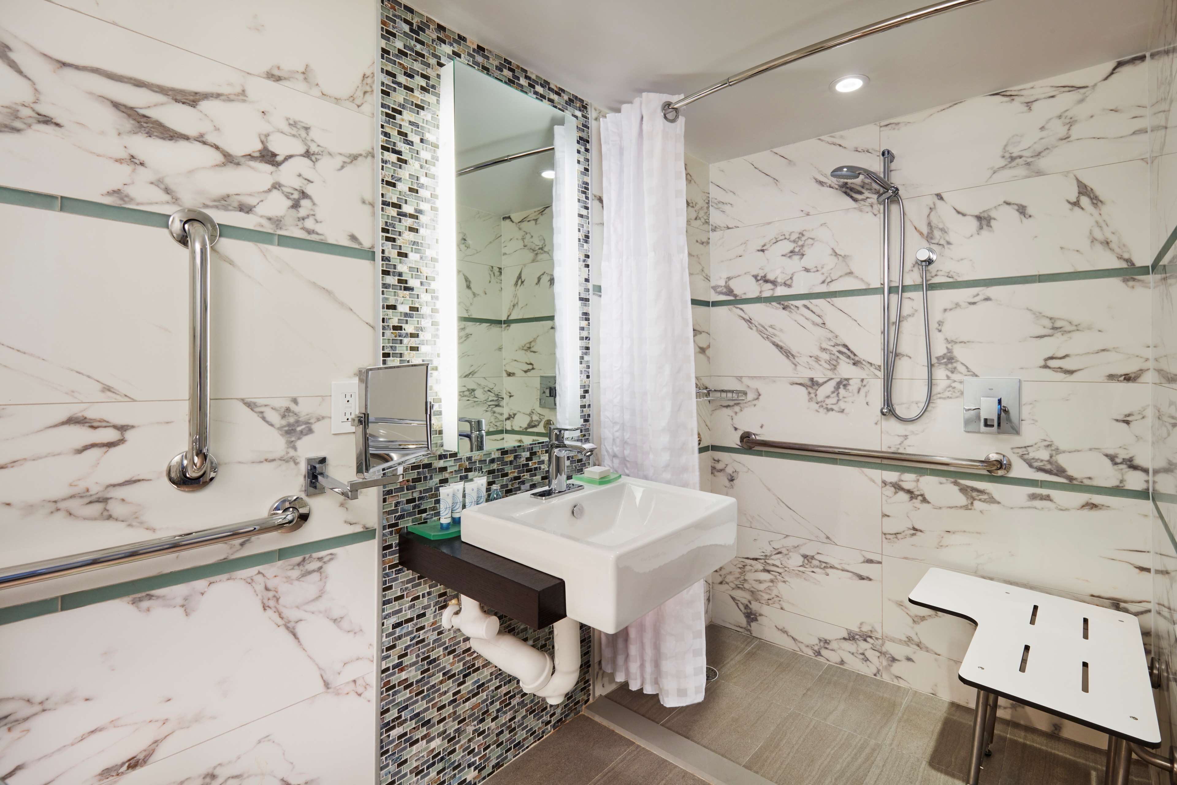 A modern bathroom with a marble backsplash, a white sink, a glass shower enclosure, and a handrail.