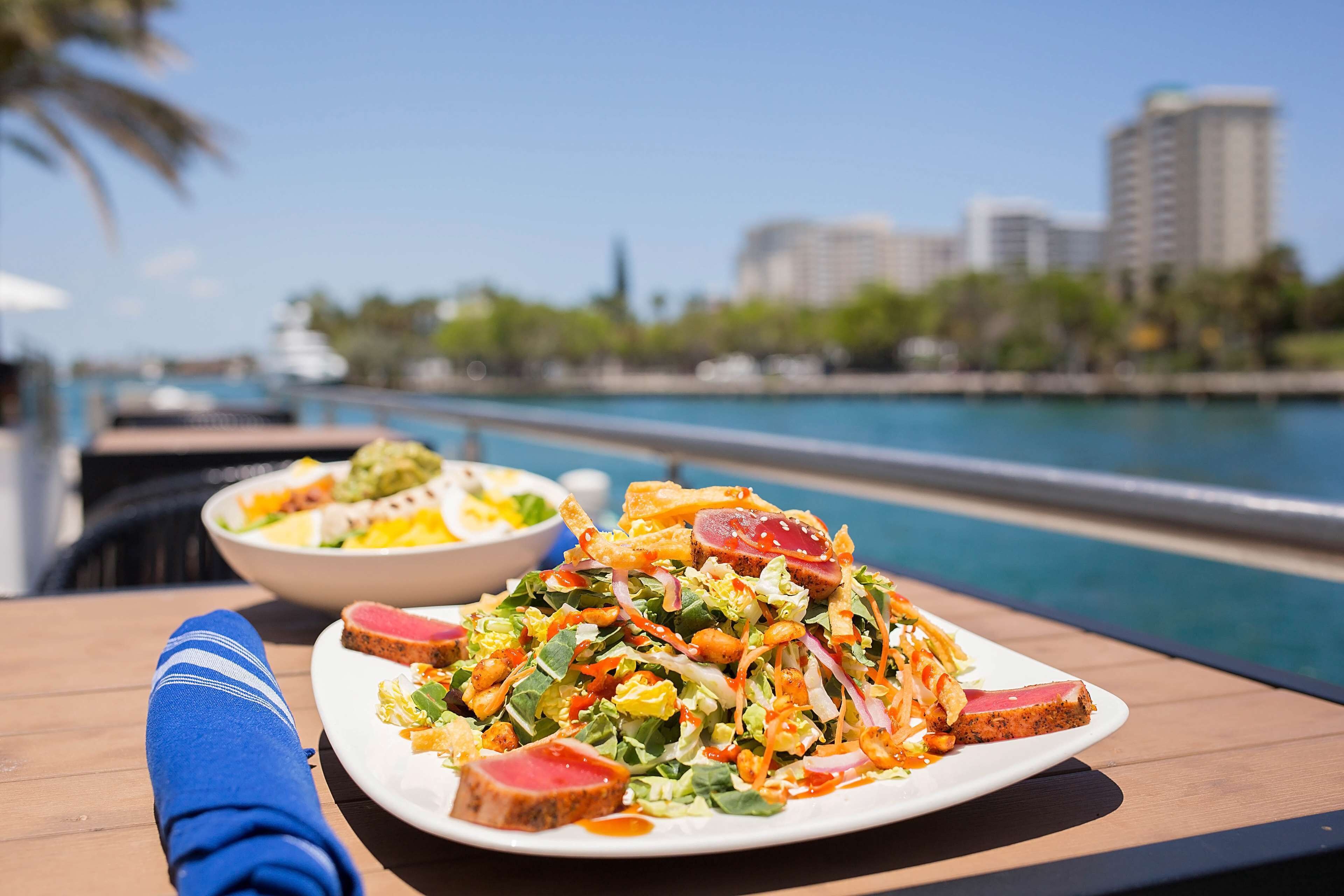 A dining table with a plate of food and a bowl of salad, overlooking a body of water with buildings in the background.