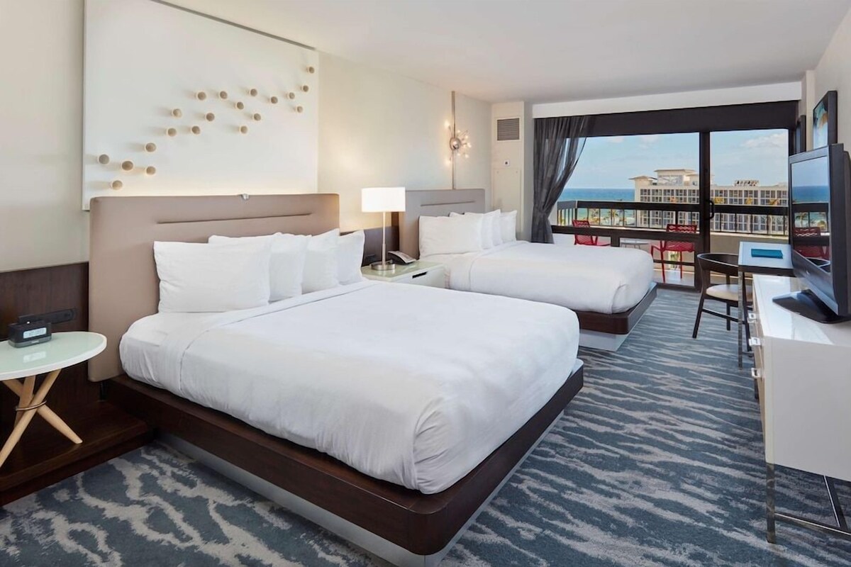A hotel room with two beds, a desk, a TV, and a view of the ocean.