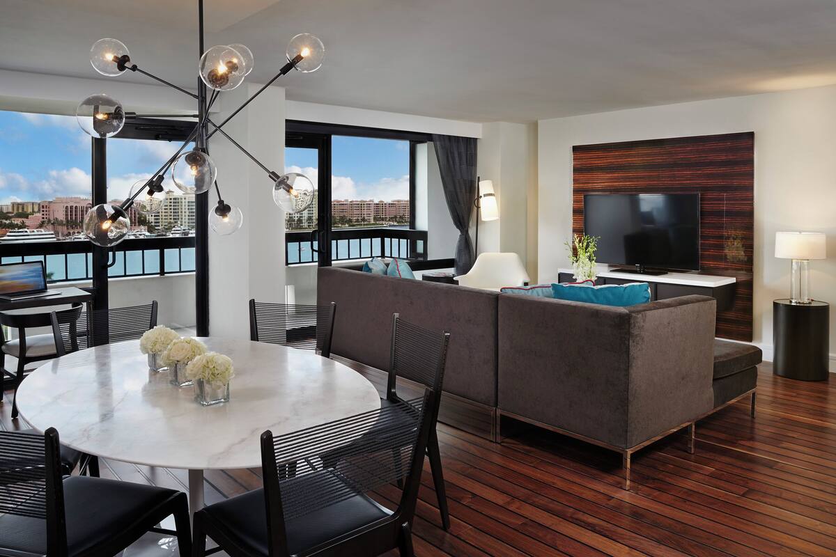 A modern hotel room with a dining area, sofa, TV, and large windows with a view.