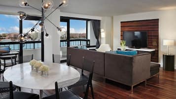 Habitación de hotel moderna con zona de comedor, sofá, televisión y ventanas amplias con vistas.