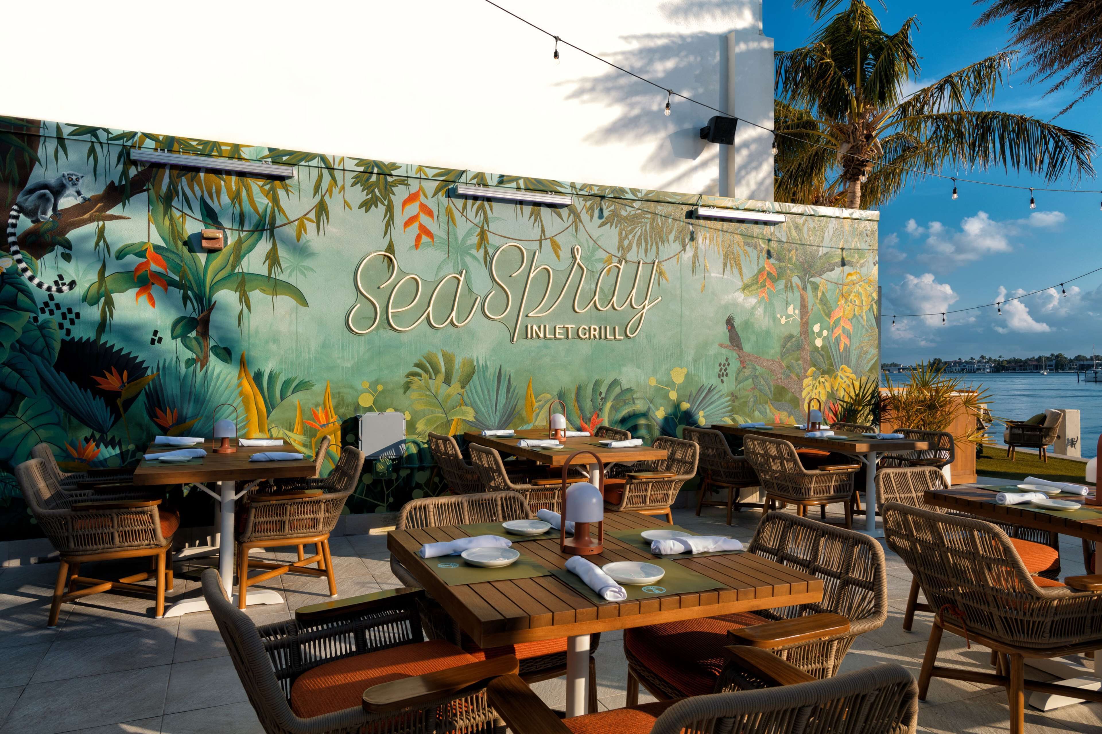 Un espace extérieur aménagé avec des tables et des chaises en osier, une fresque représentant des plantes tropicales, et l’inscription « SeaSpray Inlet Grill ».