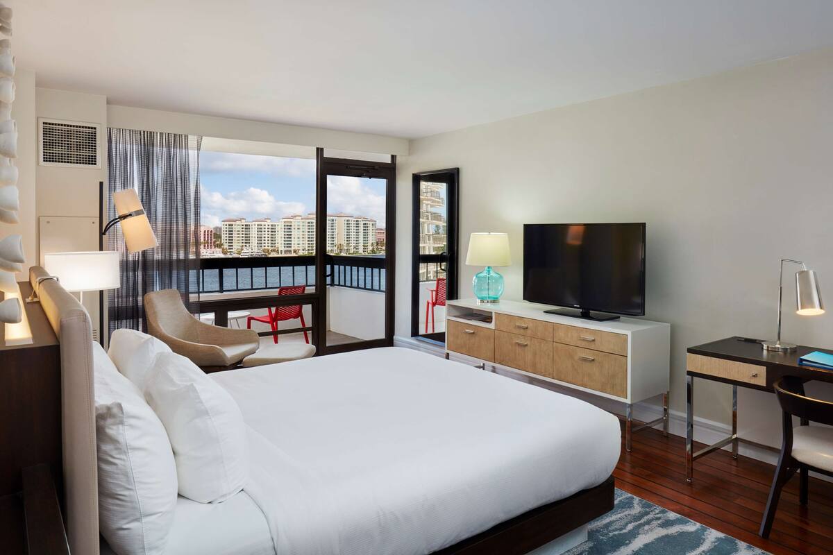 A hotel room with a large bed, a TV, a desk, and a balcony with a view of buildings.