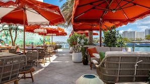 Un restaurant en bord de mer avec une terrasse, des parasols rouges, des chaises en osier et une fresque représentant un paysage tropical.