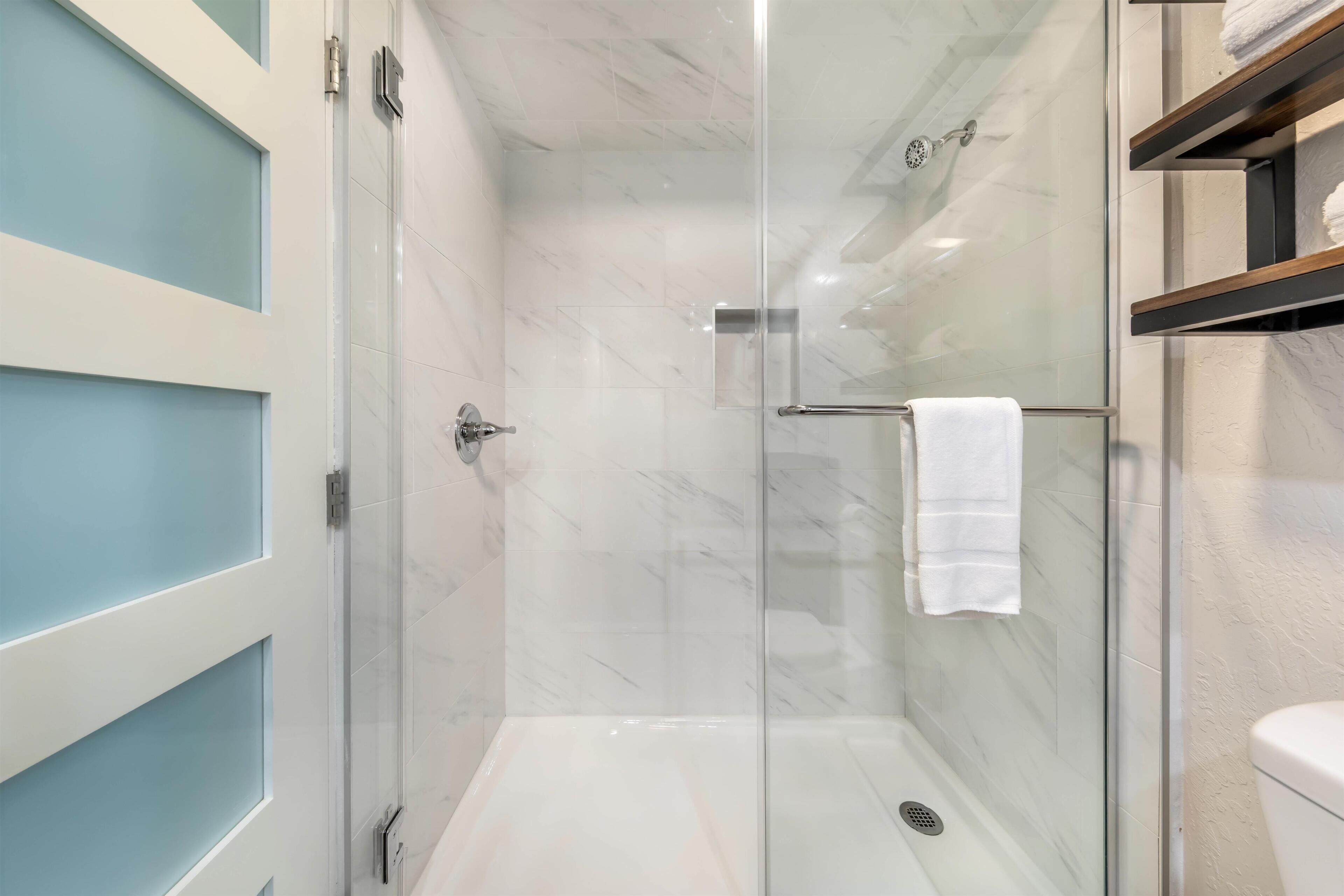 Ducha con cerramiento de vidrio, pared de mármol blanco, un perchero y un estante de madera con toallas.