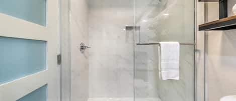 Ducha con cerramiento de vidrio, pared de mármol blanco, un perchero y un estante de madera con toallas.