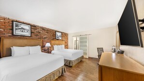 A hotel room with two beds, a wooden desk, a chair, a lamp, and a brick wall with framed pictures. - Best Western Pine Tree Motel (Chino)