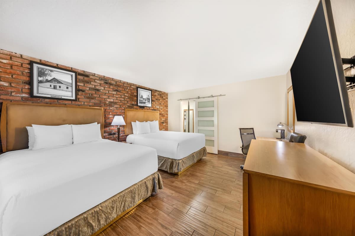 Ett hotellrum med två sängar, ett skrivbord i trä, en stol, en lampa och en vägg av tegelstenar med inramade bilder.