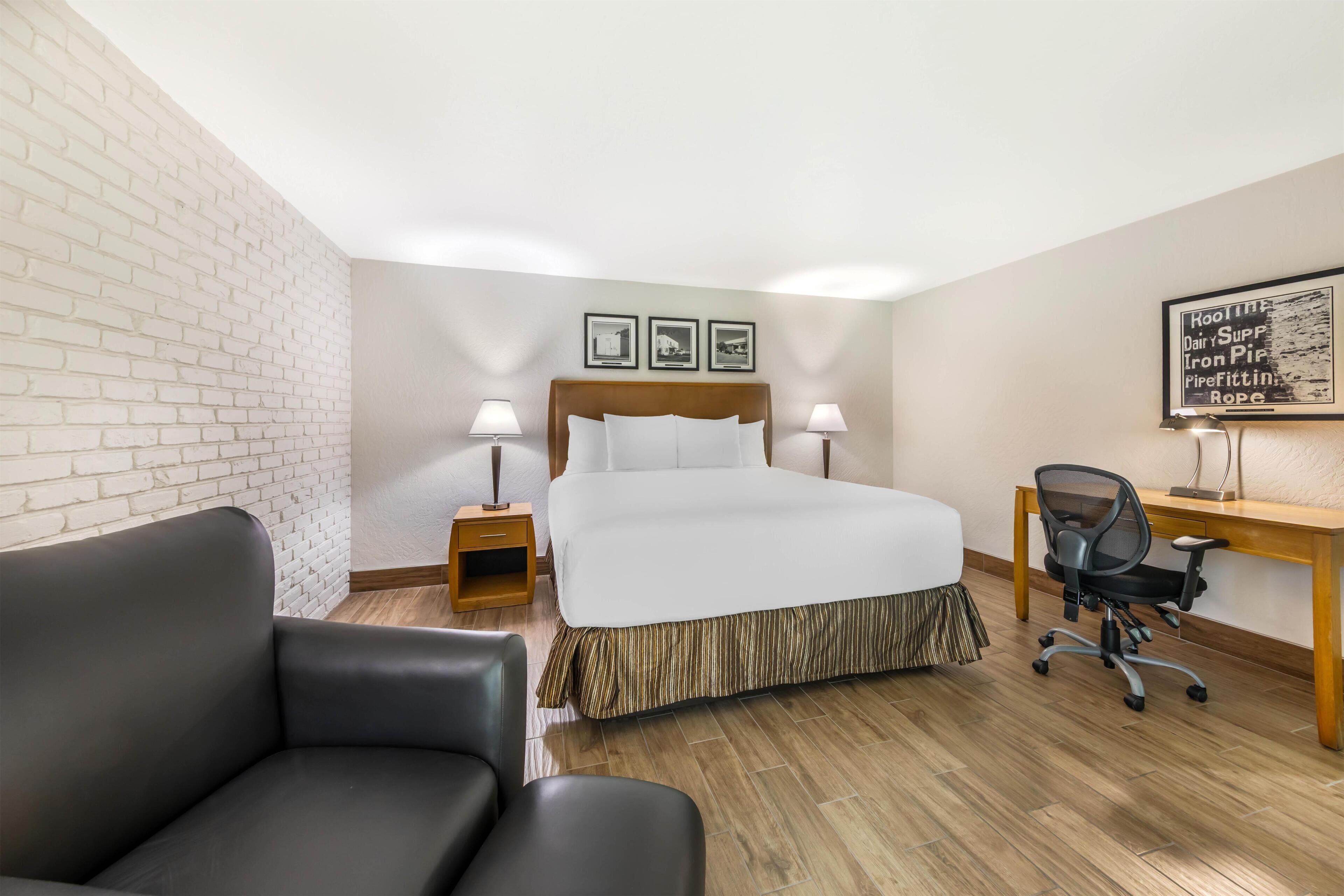 Une chambre d’hôtel avec un lit, deux lampes de chevet, un bureau avec une chaise, un canapé en cuir noir et un mur en briques.