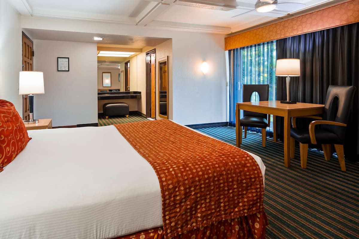 Ett hotellrum med en säng, ett skrivbord, en stol, en lampa och utsikt över korridoren.