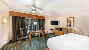 A hotel room with a bed, a desk with two chairs, a television, a mirror, and a ceiling fan.