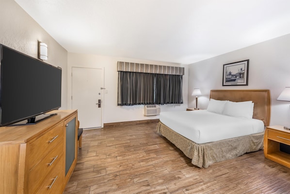 A hotel room with a bed, a TV on a wooden cabinet, a bedside table with a lamp, a window with curtains, and a picture on the wall.