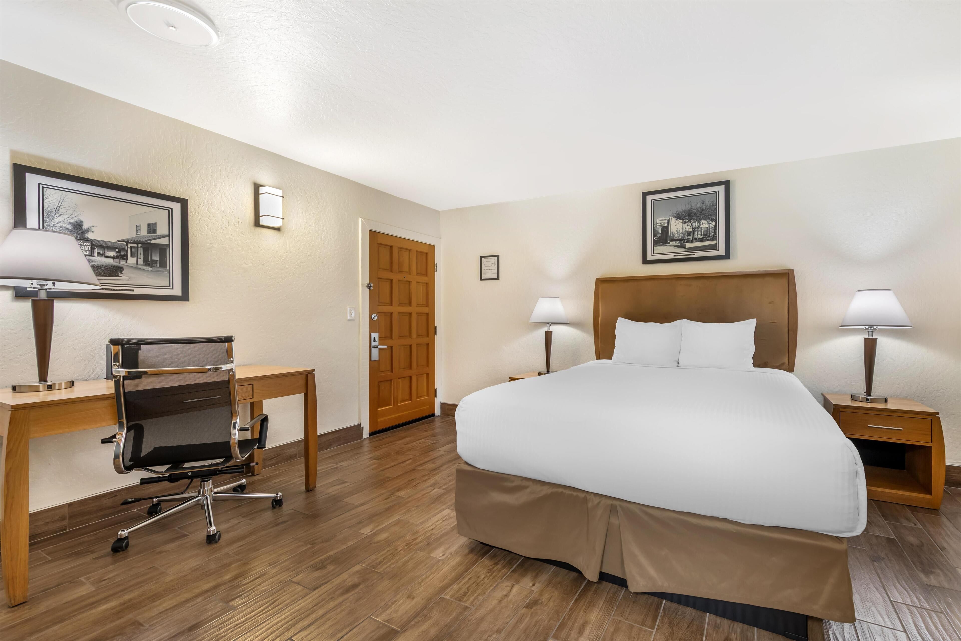 Quarto de hotel com cama, escrivaninha, cadeira, duas luminárias de cabeceira e duas imagens emolduradas nas paredes.