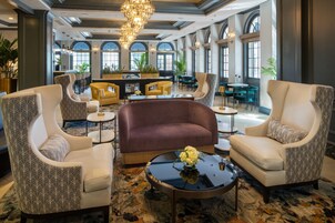 A spacious lobby with a central seating area featuring a sofa and armchairs, a bar with stools, and large windows providing natural light.