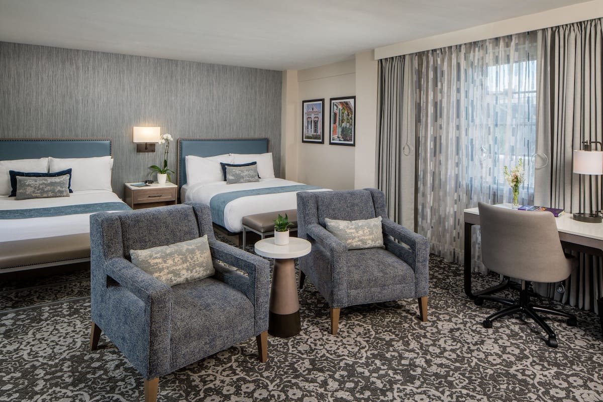 Quarto de hotel com duas camas, uma escrivaninha, uma cadeira e uma mesinha de centro com uma planta.