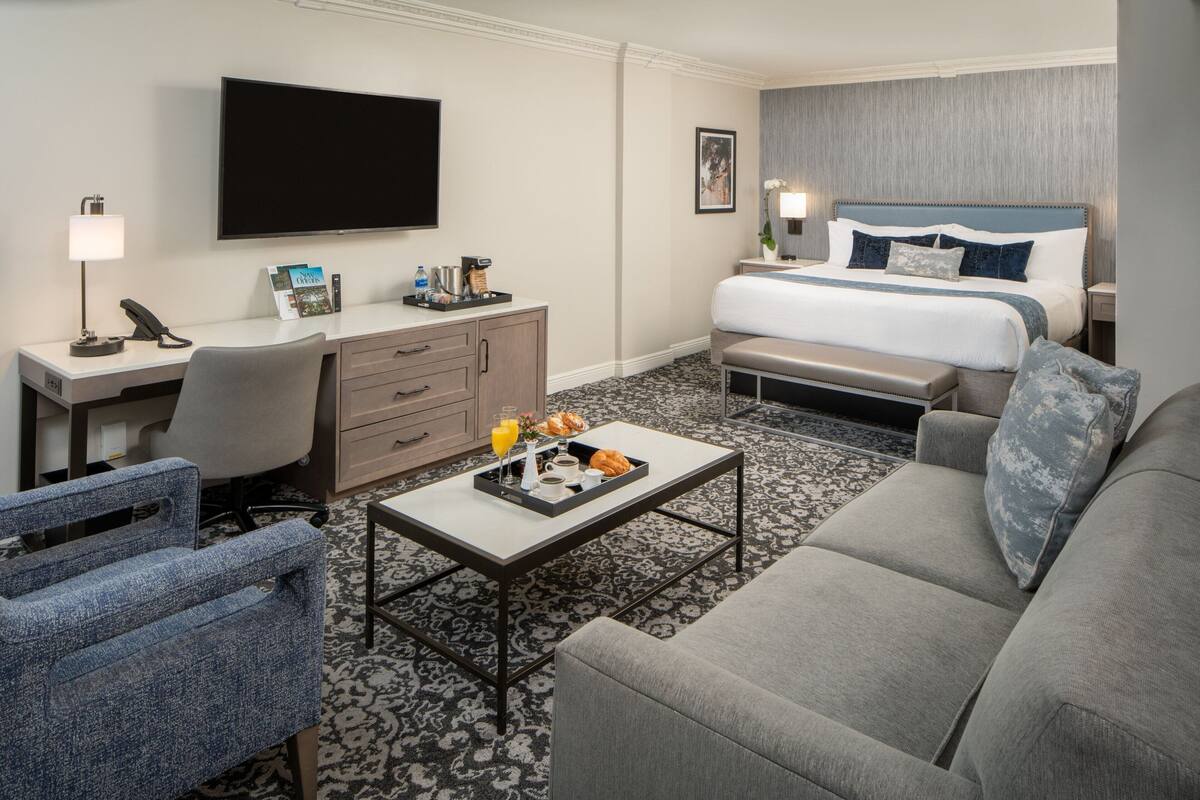 Quarto de hotel com cama, escrivaninha, cadeira, mesa de centro e televisão.