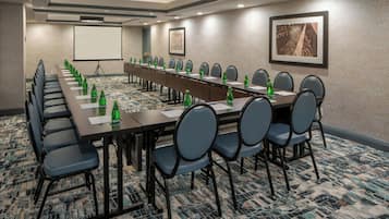 Sala de conferências com uma mesa comprida, cadeiras, tela de projetor e piso coberto por carpete.