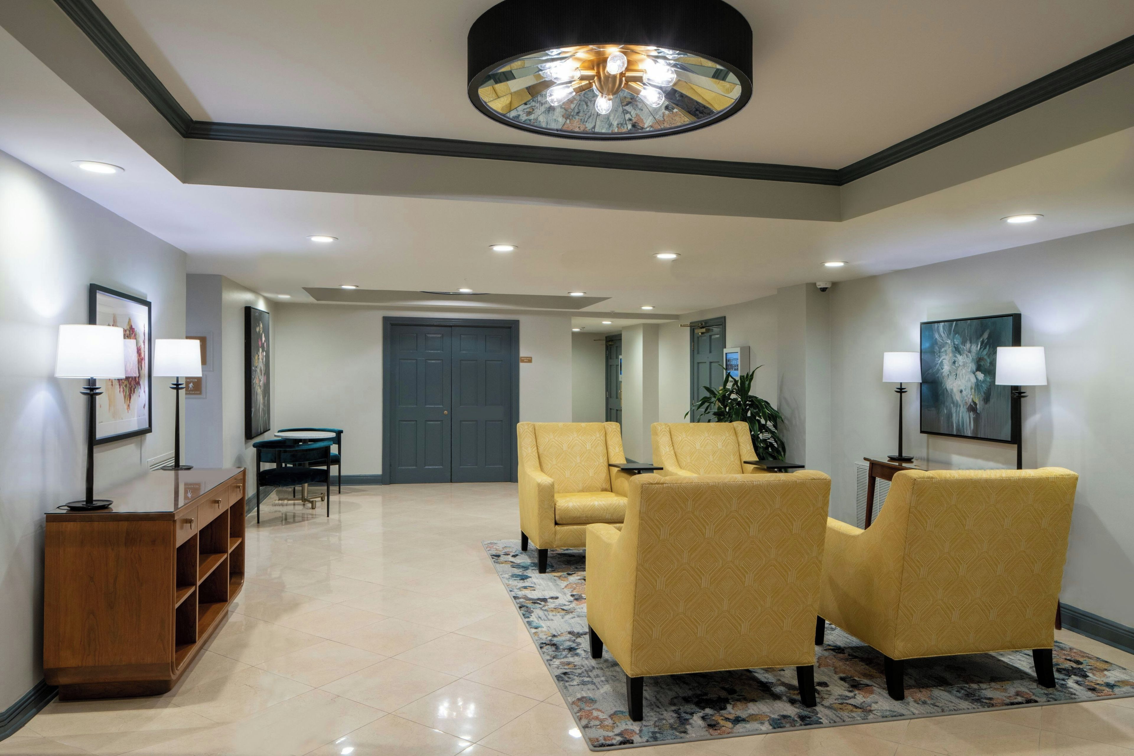 A hotel lobby with a reception desk, two armchairs, a coffee table, a large painting, and a ceiling light fixture.