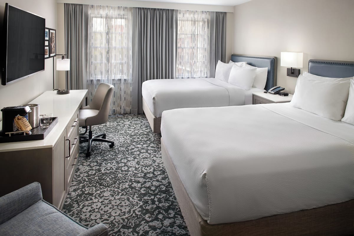 Quarto de hotel com duas camas, uma escrivaninha, uma cadeira, uma televisão e uma janela com cortinas.