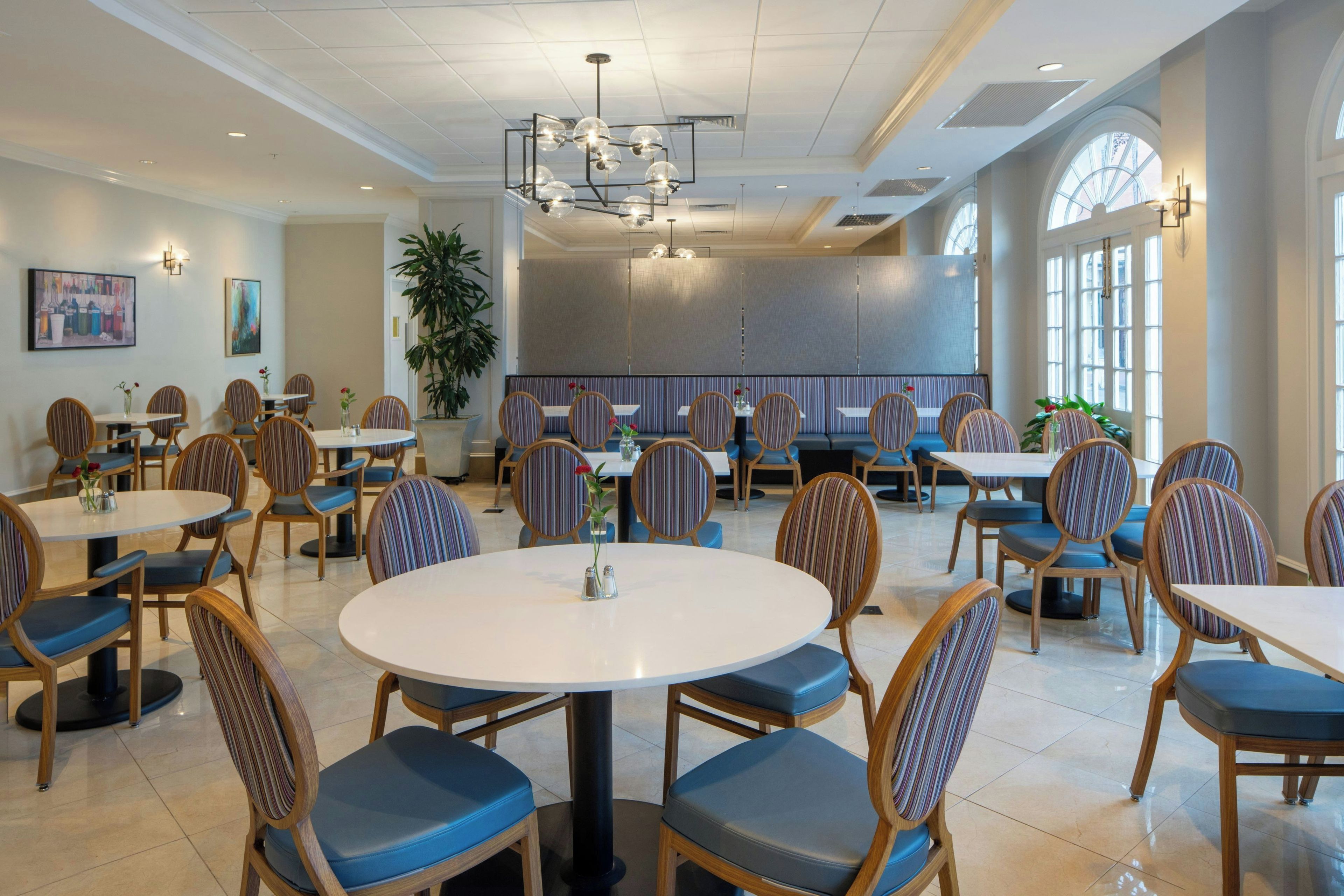 A restaurant with round tables and striped chairs, a large potted plant, and artwork on the walls.