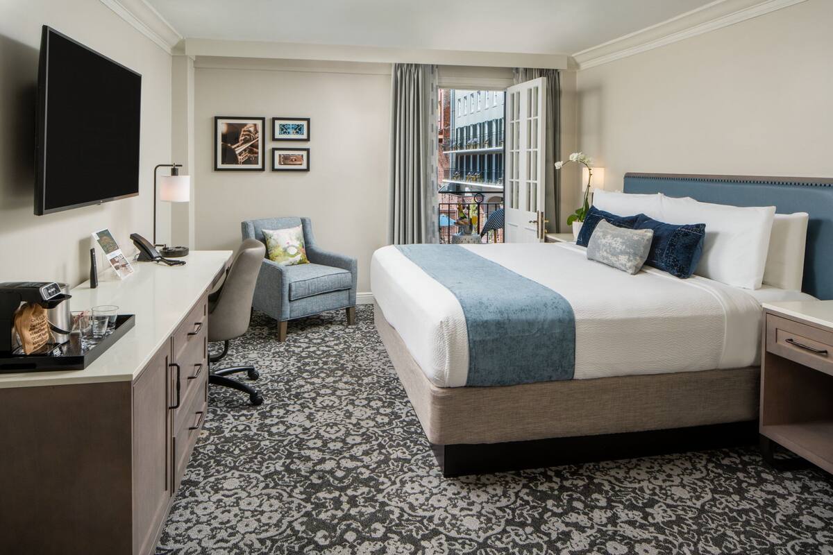 Quarto de hotel com uma cama grande, uma mesa de trabalho com telefone, uma cadeira e uma televisão fixada na parede.